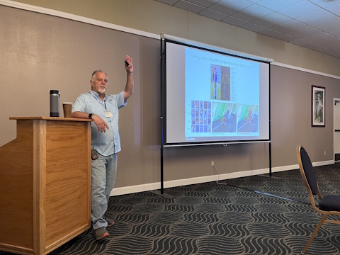 A man raises his left hand in the air, resting his right arm on a podium, while speaking beside a projector screen.